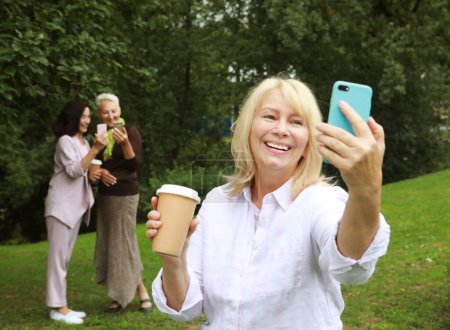 Photo for An elderly blond lady takes a selfie, holding a cup of coffee to go, in the background her female friends also use smartphones. Sunny day in the summer park. Golden Age of Retirement. - Royalty Free Image