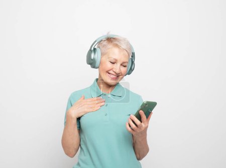 Foto de Encantador pensionista con auriculares inalámbricos. Una anciana moderna escucha música con un smartphone. - Imagen libre de derechos