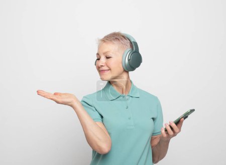 Photo for Lovely pensioner female in wireless headphones. A modern senior woman holds a smartphone and shows an empty palm presenting a product. - Royalty Free Image