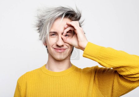 Photo for Young man wearing yellow sweater looks through zero or ok gesture, holds rounded fingers near eye - Royalty Free Image