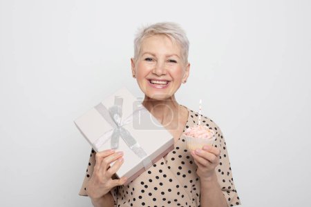 Photo for Charming pensioner woman with short haircut holding a gift and a cupcake with a candle, celebrating a birthday - Royalty Free Image