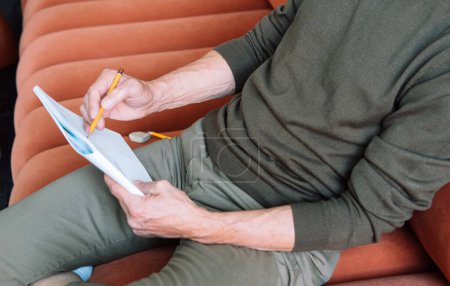 Téléchargez les photos : Homme habillé avec désinvolture assis sur le canapé et fait des croquis dans un carnet. Gros plan. - en image libre de droit