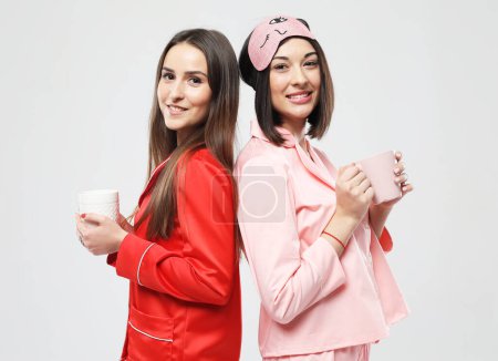 Téléchargez les photos : Deux belles jeunes femmes vêtues de pyjamas tenant des tasses sur fond gris - en image libre de droit