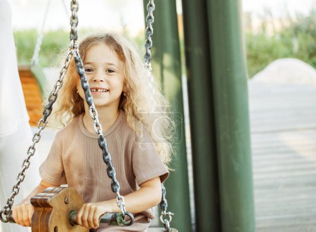 Téléchargez les photos : Fille de six ans joyeuse monte sur une balançoire dans un parc d'été. Concept de style de vie. - en image libre de droit