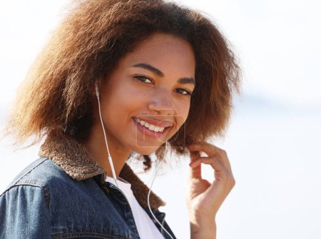 Foto de Mujer afroamericana joven escuchando música en auriculares en su teléfono móvil cerca del mar. Concepto de estilo de vida. - Imagen libre de derechos