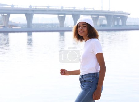 Foto de Mujer afroamericana joven con sombrero blanco escuchando música en auriculares en su teléfono móvil cerca del mar. Concepto de estilo de vida. - Imagen libre de derechos