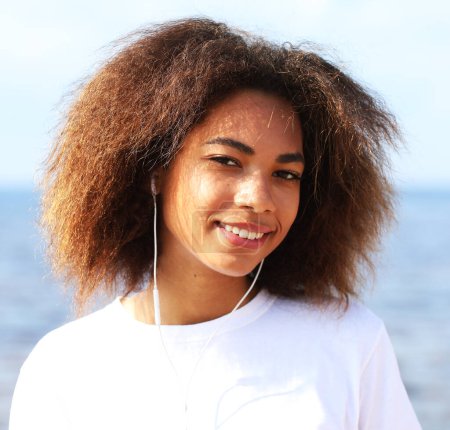 Foto de Mujer afroamericana joven escuchando música en auriculares en su teléfono móvil cerca del mar. Concepto de estilo de vida. - Imagen libre de derechos