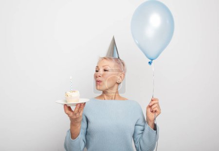 Photo for People, party, aging and maturity concept. Beauitful middle aged female wearing conical hat celebrating birthday, posing isolated with cupcake with candle and blue balloon in her hands. Happy time. - Royalty Free Image