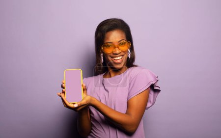 Téléchargez les photos : Portrait de jeune promotrice afro-américaine positive montrer smartphone sur fond violet - en image libre de droit