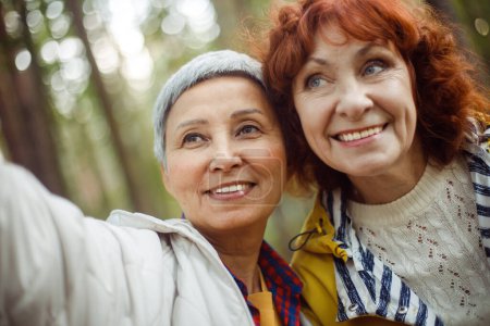 Téléchargez les photos : Femmes âgées multiraciales s'amuser pendant la journée de trekking dans le bois et faire du selfie avec smartphone. Parc d'automne. - en image libre de droit