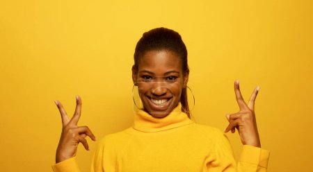 Photo for Young smiling black woman 20s wears yellow sweater showing covering with victory sign - Royalty Free Image