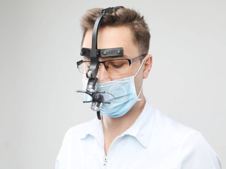 Foto de Médico varón caucásico con una máscara quirúrgica y lupas binoculares en el fondo gris, de cerca - Imagen libre de derechos