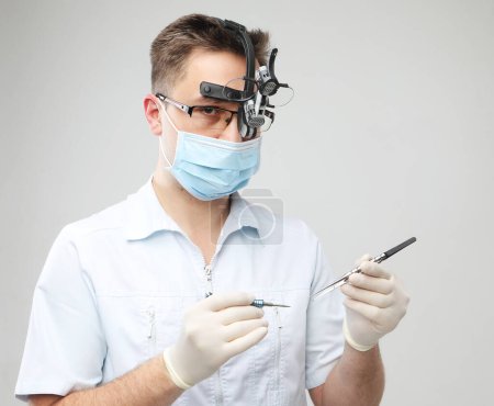 Téléchargez les photos : Médecin homme caucasien dans un masque chirurgical et loupes binoculaires tenant des instruments dentaires à l'arrière-plan gris, gros plan - en image libre de droit