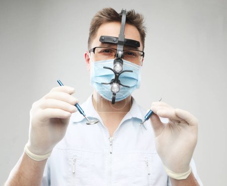 Foto de Médico varón caucásico con máscara quirúrgica y lupas binoculares sosteniendo instrumentos dentales en el fondo gris, de cerca - Imagen libre de derechos