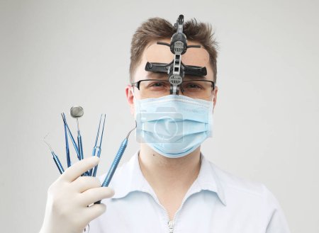 Foto de Médico varón caucásico con máscara quirúrgica y lupas binoculares sosteniendo instrumentos dentales en el fondo gris, de cerca - Imagen libre de derechos