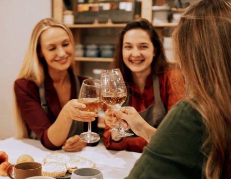 Foto de Tres alegres amigas jóvenes se están divirtiendo en un taller de cerámica. Beben vino y bromean, se divierten. Tiempo de fiesta. - Imagen libre de derechos