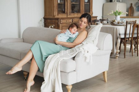 Foto de Una joven madre feliz está sentada en el sofá y sosteniendo a un bebé en sus brazos. Concepto de infancia y familia. - Imagen libre de derechos