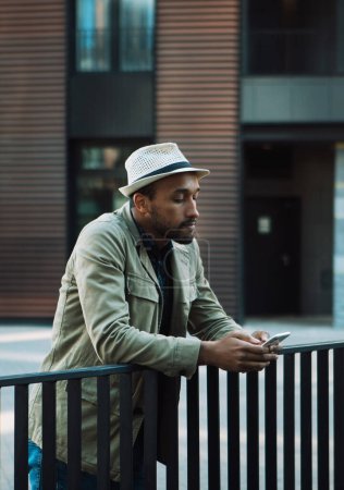 Foto de Joven Afro hombre con gafas de sol usando smartphone a mensaje de texto cerca del moderno centro de negocios al aire libre. Concepto de estilo de vida y personas. - Imagen libre de derechos