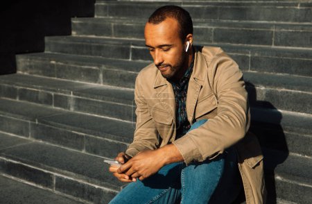 Téléchargez les photos : Portrait de jeune homme afro-américain assis sur les marches à l'aide d'un téléphone portable. - en image libre de droit