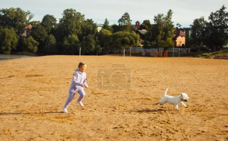 Téléchargez les photos : A seven-year-old girl with pigtails plays with a west highland white terrier on the beach near the lake. Lifestyle concept. - en image libre de droit