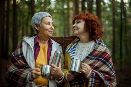 Two elderly women friends walk in the forest, pour coffee from a thermos, have a great time together. Lifestyle and people concept.