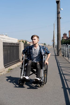 Foto de Un joven discapacitado cabalga en una silla de ruedas a través de un puente, el joven hombre vestido con una camisa a cuadros y vaqueros. Concepto de rehabilitación, salud y apoyo a las personas con discapacidad. - Imagen libre de derechos