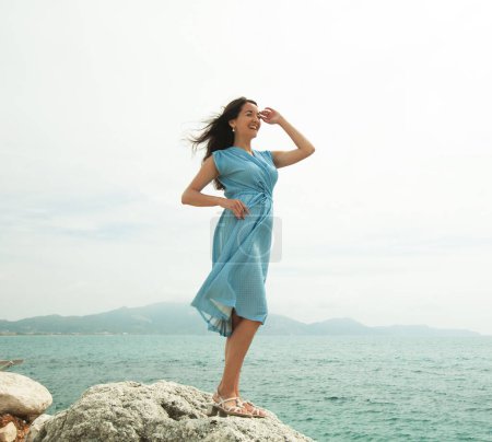 Téléchargez les photos : Jeune femme heureuse portant une robe bleue posant près de la mer, vacances d'été - en image libre de droit