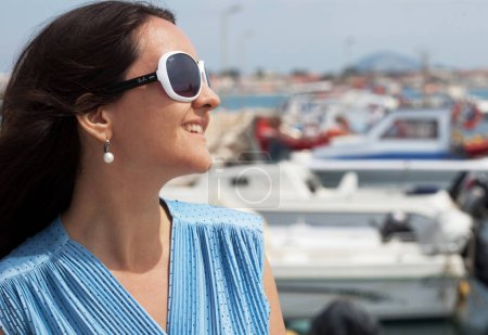 Téléchargez les photos : Belle jeune femme brune en robe bleue et chapeau se tient au port et regarde les bateaux - en image libre de droit