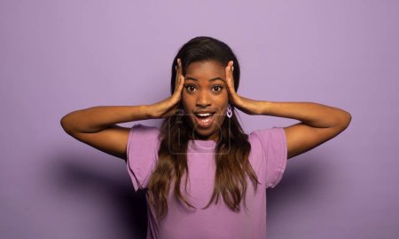 Téléchargez les photos : Jeune femme afro-américaine est étonnée avec sa bouche ouverte, regarde loin dans la surprise. Portrait sur fond violet. - en image libre de droit