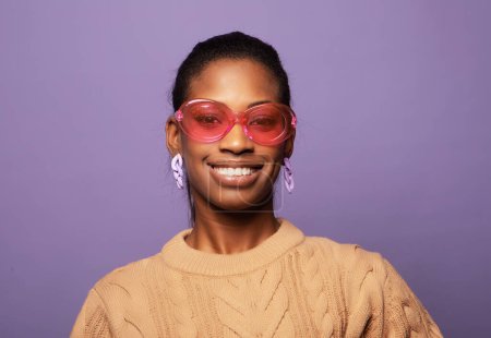 Téléchargez les photos : Charmante jeune femme noire à la mode des années 20 porte un pull beige et des lunettes de soleil roses sur fond violet. - en image libre de droit