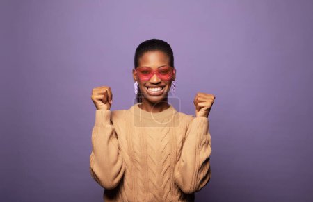 Téléchargez les photos : Jeune femme afro-américaine portant un pull et des lunettes de soleil célèbre la victoire et le succès, se sent vif et énergique sur fond violet - en image libre de droit