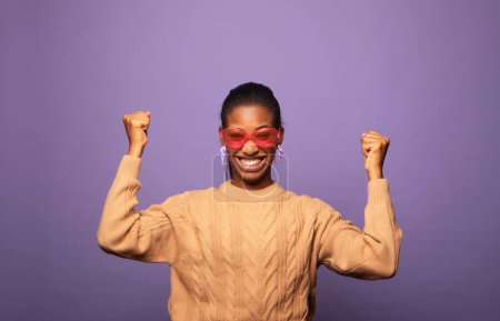 Photo for Young African American woman wearing sweater and sunglasses celebrates victory and success, feels lively and energetic over purple background - Royalty Free Image