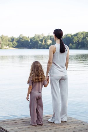 Foto de Una joven madre y su hija de cinco años observan los peces en el lago, de pie en el puente. Vista trasera. Concepto de familia y personas. - Imagen libre de derechos