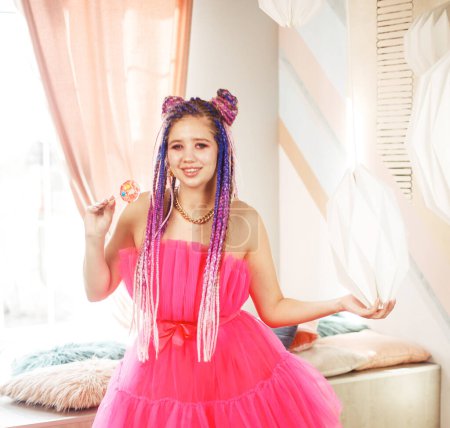 Téléchargez les photos : Jeune femme heureuse avec dreadlocks coiffure portant robe rose, style poupée, maquillage lumineux, dans la chambre rose. - en image libre de droit