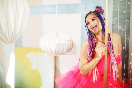 Foto de Mujer feliz joven con rastas peinado con vestido rosa, estilo muñeca, maquillaje brillante, hasta en la habitación rosa. - Imagen libre de derechos