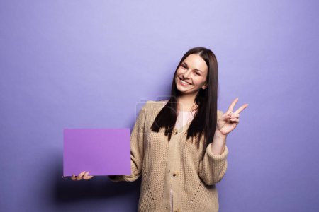 Photo for Young smiling woman holding purple blank advertising board standing on lilac background in studio - Royalty Free Image