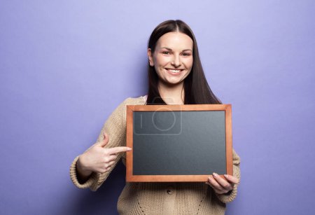 Photo for Young smiling woman holding a chalkboard isolated on purple background. - Royalty Free Image