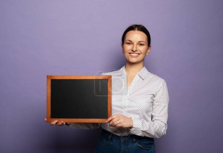 Téléchargez les photos : Young smiling woman holding a chalkboard isolated on purple background. - en image libre de droit