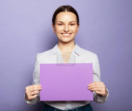 Téléchargez les photos : Jeune femme souriante tenant le panneau publicitaire vierge violet debout sur fond lilas en studio - en image libre de droit