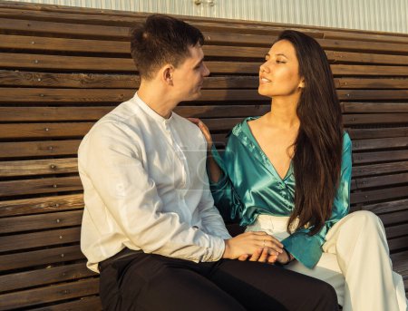 Téléchargez les photos : Un jeune couple amoureux de nationalités différentes s'assoit sur un banc et admire le coucher de soleil. Bonne heure d'été. - en image libre de droit