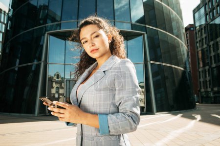 Photo for A young african woman dressed in business style against the background of a business center is holding a mobile phone - Royalty Free Image