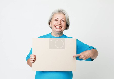 Foto de Una anciana feliz y encantadora sostiene una hoja de papel en blanco en sus manos y la señala con su dedo. Aislado sobre fondo blanco. - Imagen libre de derechos