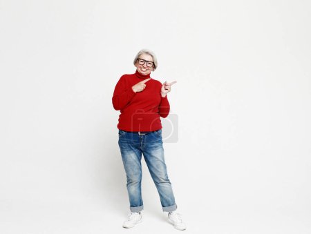 Foto de Smiling elderly gray-haired blonde woman lady 60s years old in red casual sweater pointing index finger aside up on mock up copy space isolated on white background studio portrait - Imagen libre de derechos