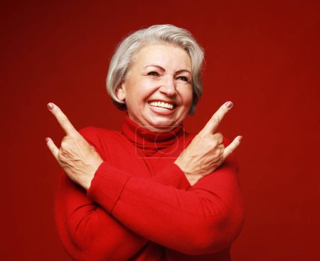Téléchargez les photos : Femme âgée rire et montrant signe de roche à la caméra sur fond rouge. Émotion et sentiments. - en image libre de droit