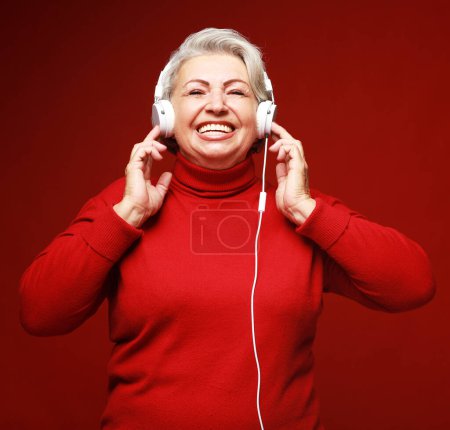 Photo for Lifestyle, party, emotion and old people concept: elderly woman wearing red sweater listening to music with headphones - Royalty Free Image