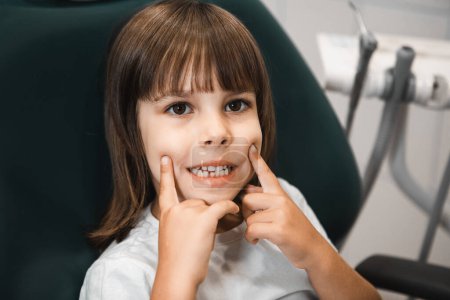 Photo for A little girl at a dentist appointment, smiles and touches her cheeks showing a smile - Royalty Free Image