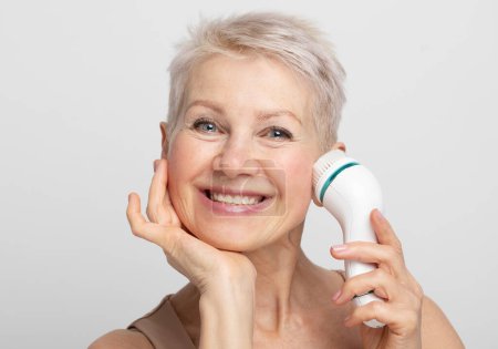 Photo for Beauty face portrait of cute and pretty senior woman with short hair using facial massager for perfect fresh skin over grey background - Royalty Free Image