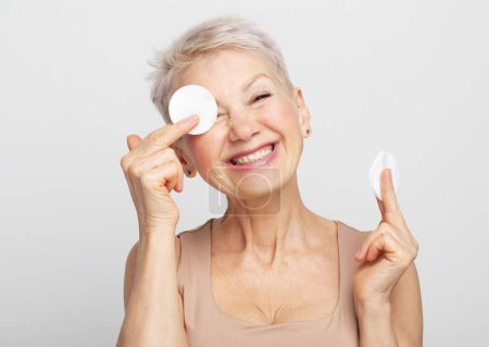 Foto de Hermosa anciana de 60 años mujer con pelo corto haciendo su rutina diaria quitándose el maquillaje con algodón - Imagen libre de derechos