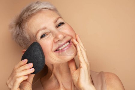 Téléchargez les photos : Photo de femme âgée avec des cheveux courts utiliser éponge enlever le maquillage sur fond beige, gros plan - en image libre de droit