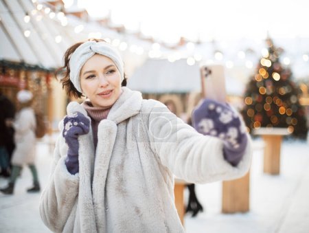 Photo for Young smiling woman in winter clothes at the Christmas market takes a selfie on a smartphone - Royalty Free Image